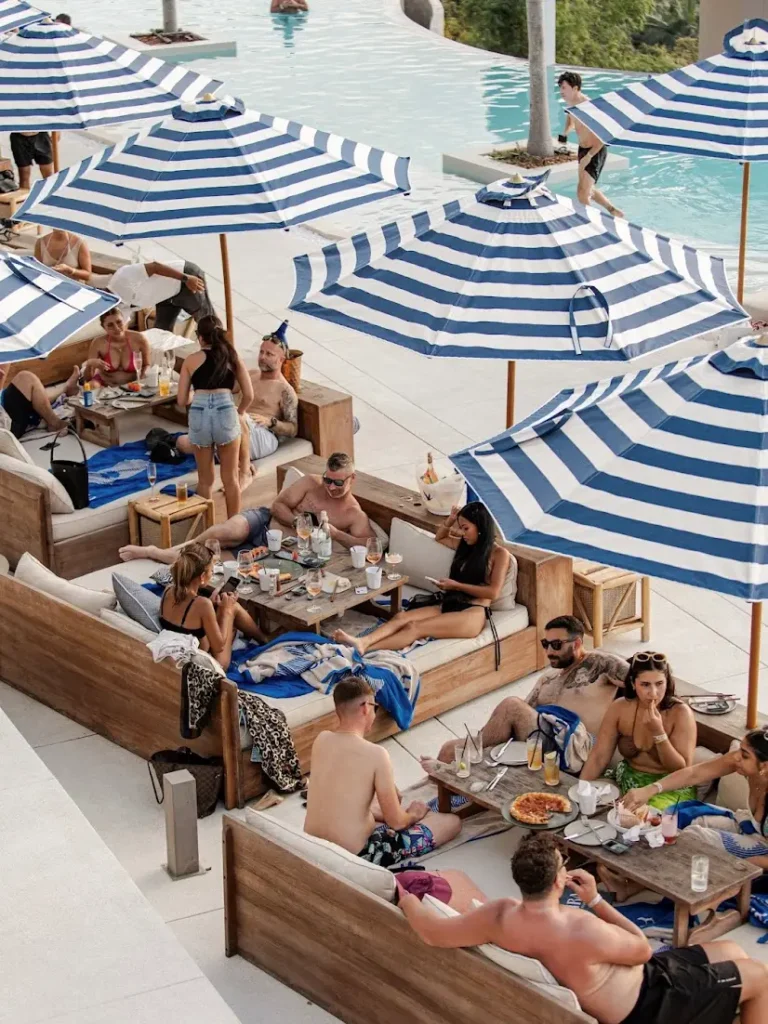 Guests relaxing on contemporary outdoor loungers, sofas, and daybeds made from recycled teak wood at Fira Beach Club, Phuket, with striped umbrellas and a poolside view